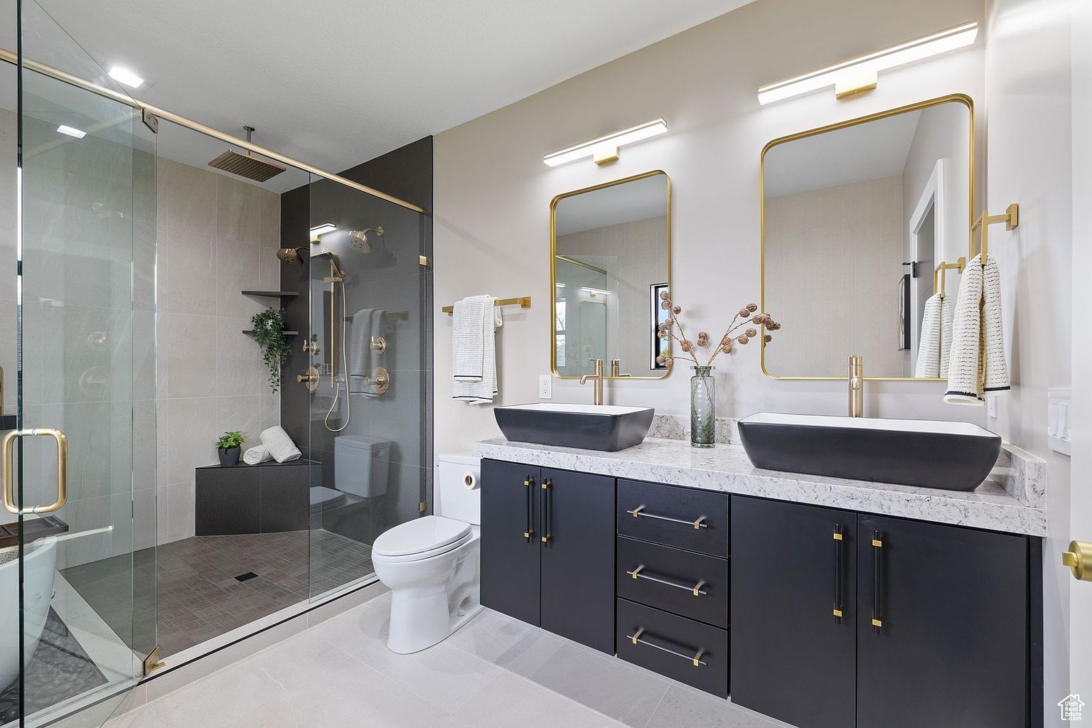 This is a modern primary bathroom featuring a glass-enclosed shower with gold fixtures and a built-in bench. The vanity has dual black vessel sinks, a white countertop, and dark blue cabinetry with gold hardware. The overall design is sleek and luxurious, creating a spa-like atmosphere.