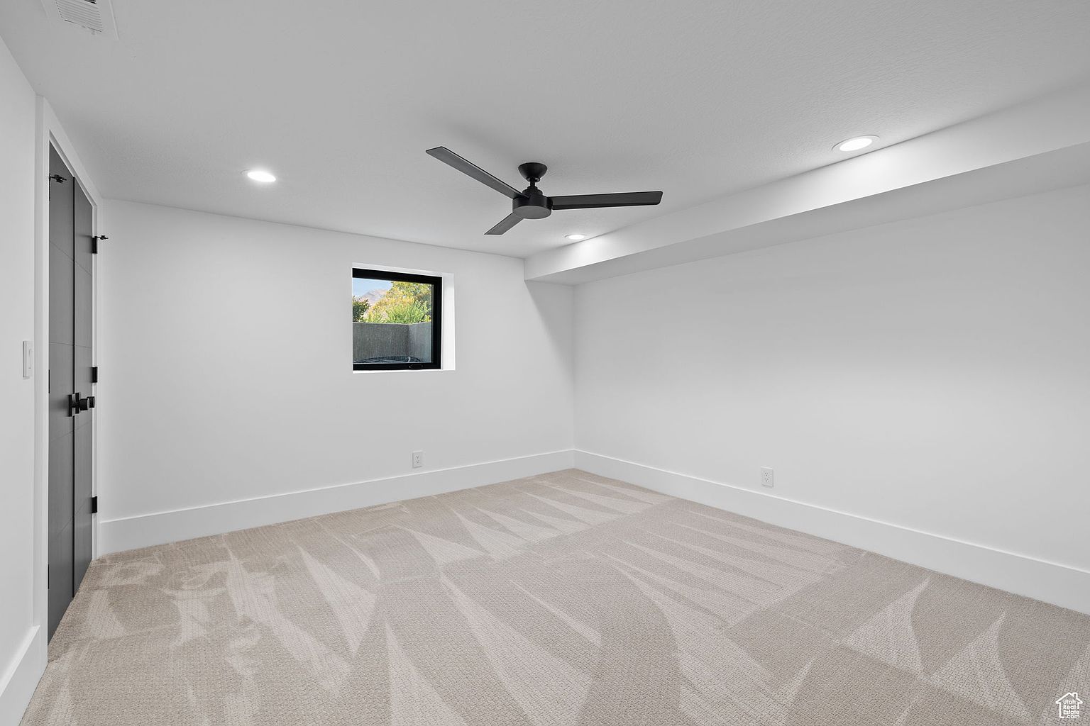 The image showcases a clean and modern bedroom with white walls and light beige carpet. A black ceiling fan adds a contemporary touch, while a window provides natural light. The room is spacious and uncluttered, presenting a blank canvas for potential buyers.