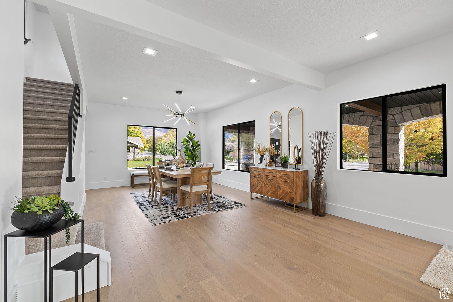 This is an interior shot of a modern dining room. The room features light wood flooring, white walls, and black-framed windows offering natural light. A dining table with chairs sits on a patterned rug, complemented by a modern chandelier and a wooden sideboard with decorative mirrors and vases. A staircase is visible on the left side of the image.