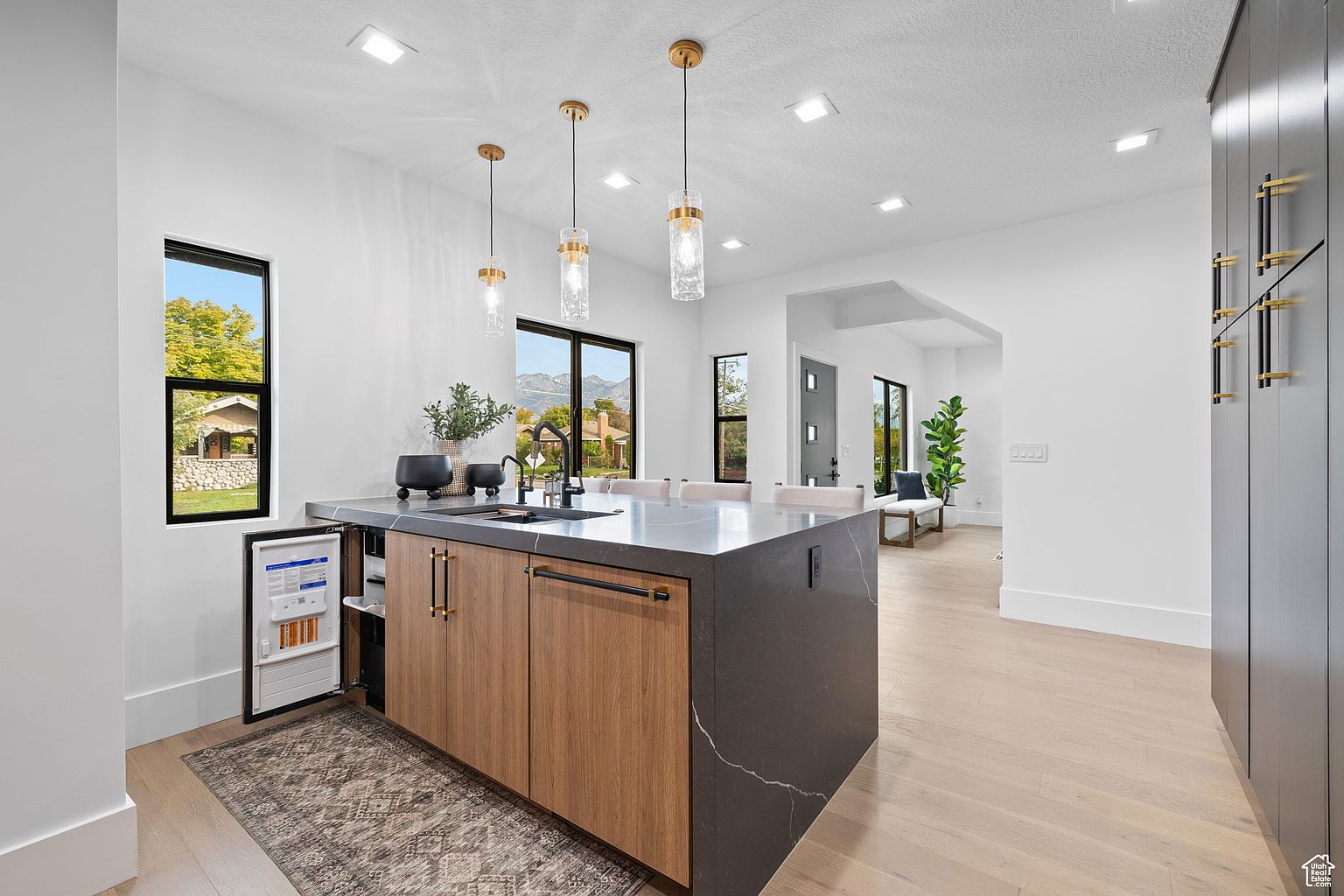 This is a bright, modern kitchen featuring a large island with a dark countertop and wood cabinetry. The island includes a built-in beverage refrigerator. Three pendant lights hang above the island, and natural light streams in from the windows, offering a view of the outdoors.