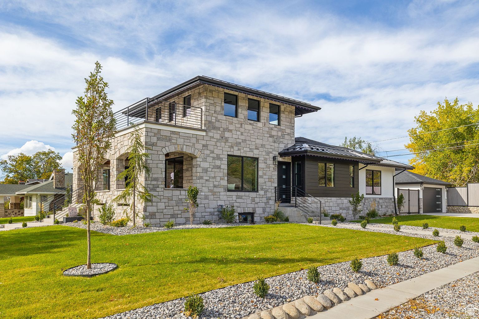 This is a front exterior view of a modern two-story home with a stone facade. The house features a balcony on the second floor, a well-manicured lawn, and a stone pathway leading to the entrance. The overall impression is one of contemporary elegance and curb appeal.