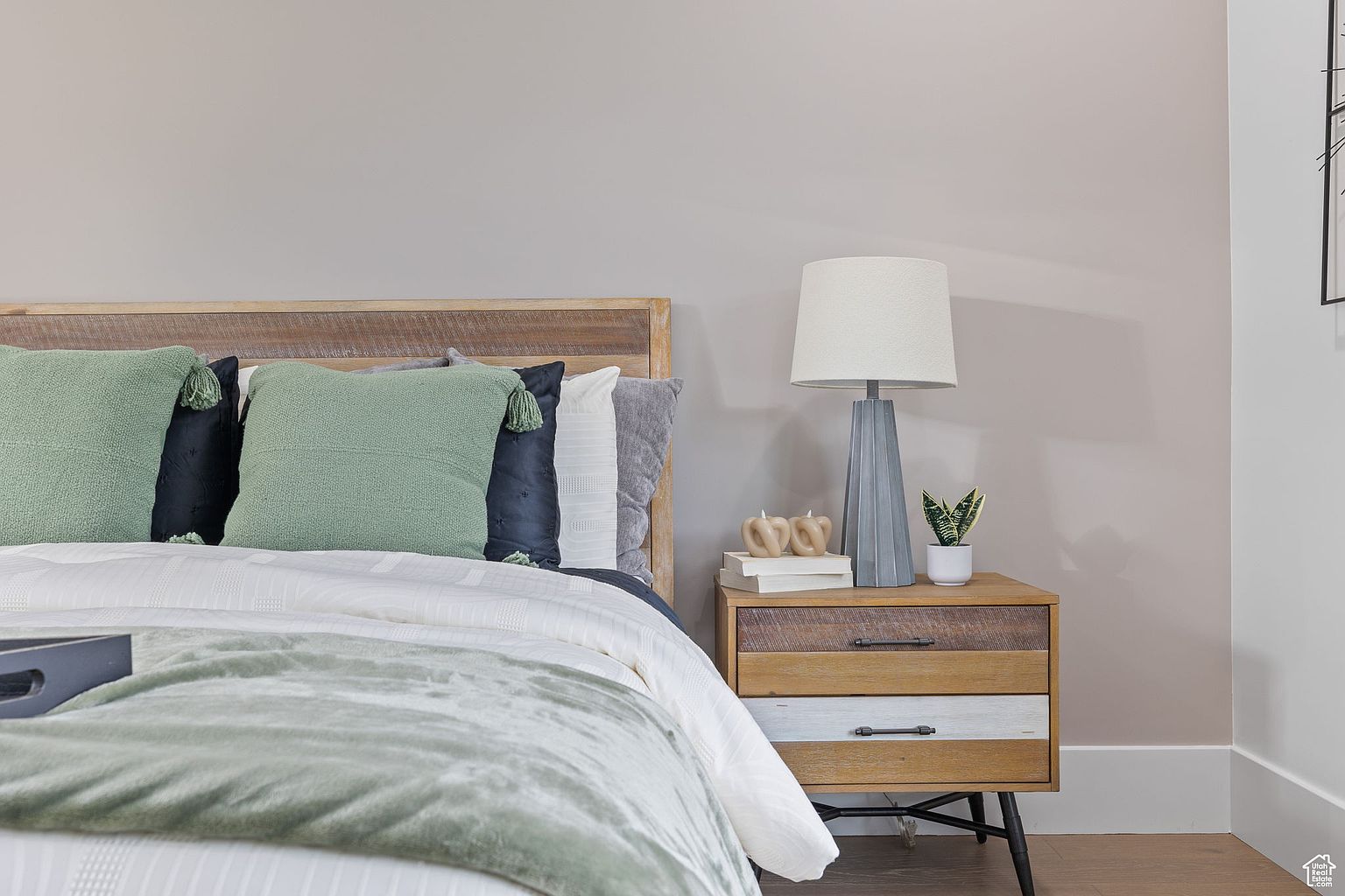 This is a cozy bedroom featuring a wooden-framed bed with green and navy pillows, and a white comforter with a sage green throw. A wooden nightstand with a gray lamp, books, candles, and a small plant sits beside the bed. The walls are a neutral beige, creating a calming and inviting atmosphere.