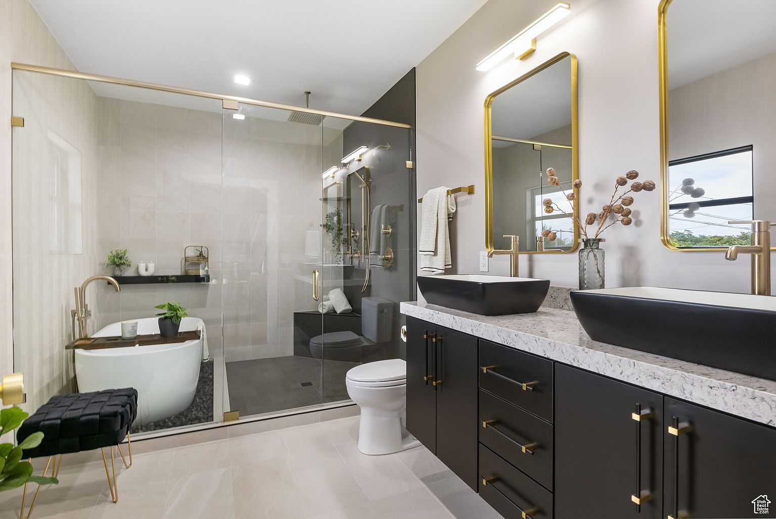 This is a luxurious primary bathroom featuring a glass-enclosed shower and soaking tub area with modern fixtures and finishes. The vanity has dual black vessel sinks, gold hardware, and a light-colored countertop, complemented by gold-framed mirrors and sconces. The overall design is contemporary and elegant, creating a spa-like atmosphere.