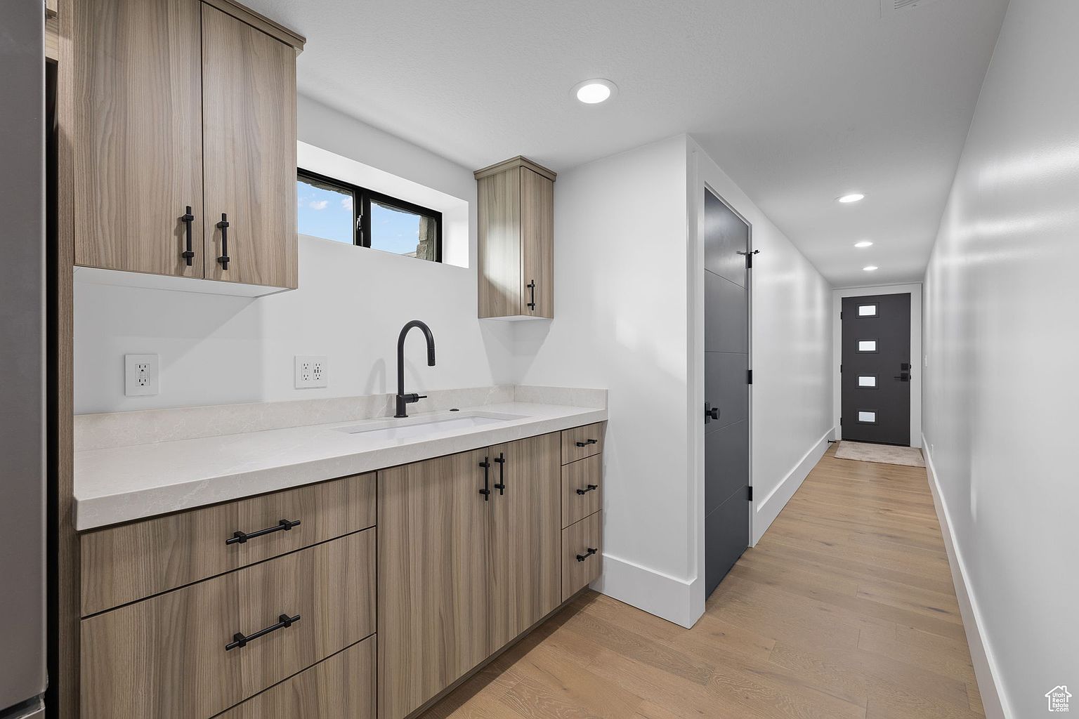 This interior shot showcases a modern kitchen area with light wood-grain cabinetry and black hardware. A sleek black faucet complements the light countertop, and a window provides natural light above the sink. The space transitions into a hallway with light wood flooring and a dark door, creating a clean and contemporary aesthetic.