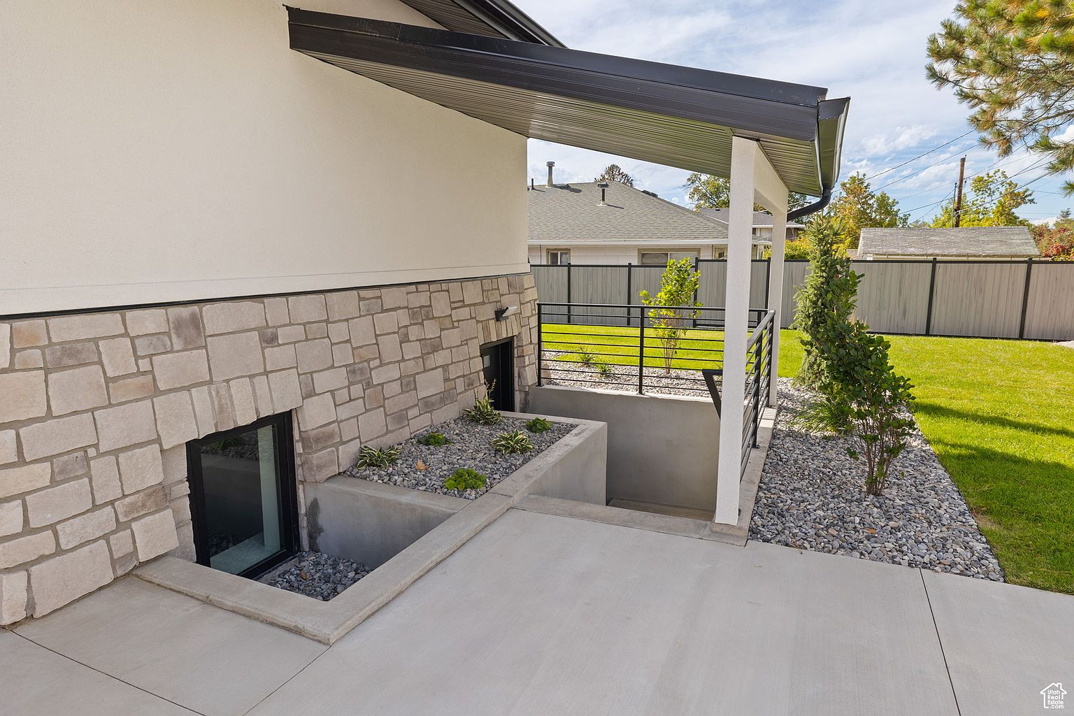 This exterior side view showcases a modern home with a combination of stucco and stone facade. A covered area with white columns provides shelter, while a well-maintained yard with gravel and greenery adds to the curb appeal. The design incorporates window wells with decorative stone and plantings, enhancing the aesthetic appeal of the basement area.