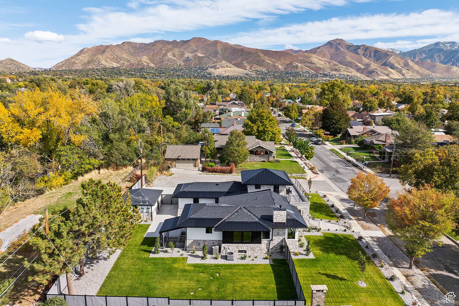 This aerial shot showcases a modern home with a dark roof and light-colored exterior, surrounded by a well-manicured lawn and mature trees. The property is situated in a neighborhood with mountain views in the background, creating a serene and desirable setting. The overall impression is one of luxury and privacy.
