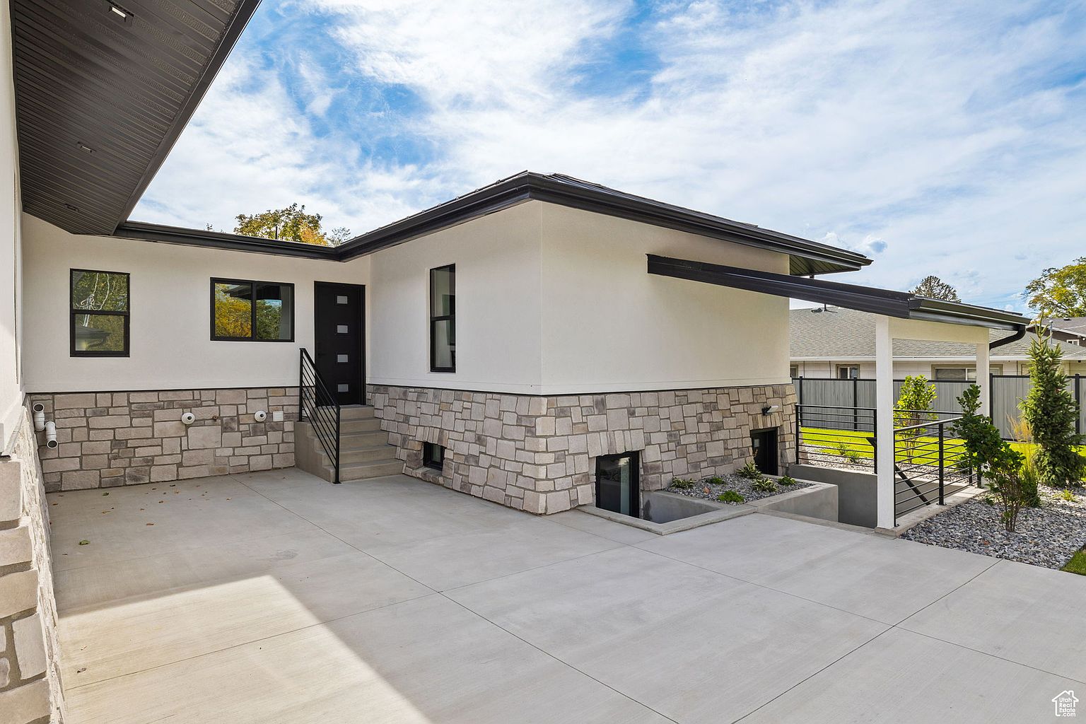 This is a side view of a modern home featuring a combination of white stucco and stone veneer on the lower level. The house has black-framed windows and a dark-colored roof. A concrete driveway leads to the side of the house, and landscaping is visible on the right side of the frame.