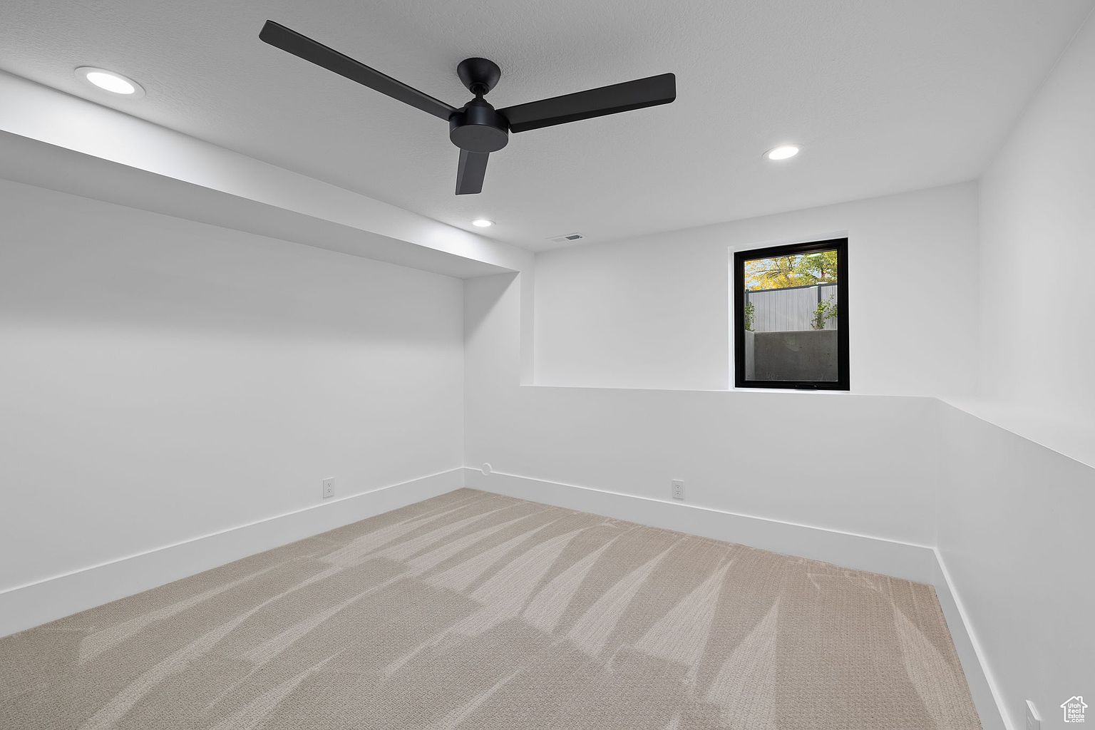 This is a clean and modern bedroom featuring neutral-toned carpet and white walls. A black ceiling fan adds a contemporary touch, while a window provides natural light. The room is simple and well-lit, creating a welcoming and versatile space.