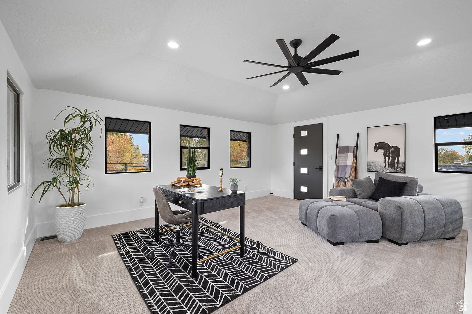 This is an interior shot of a modern office space. The room features a black desk with gold accents, a gray office chair, and a black and white patterned rug. A gray sectional sofa sits to the right, and natural light streams in through several windows, creating a bright and inviting workspace.