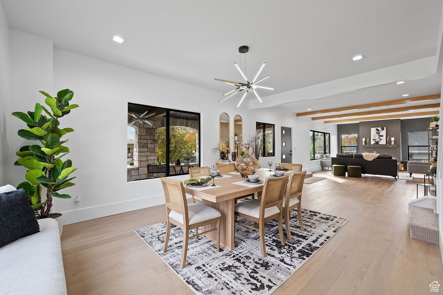 This is an interior shot of a dining room featuring a light wood dining table with six chairs, set on a black and white patterned rug. A modern chandelier hangs above the table, and natural light streams in through a large window, highlighting the open floor plan that leads into the living room. The space is decorated with neutral tones and contemporary furnishings, creating a bright and inviting atmosphere.