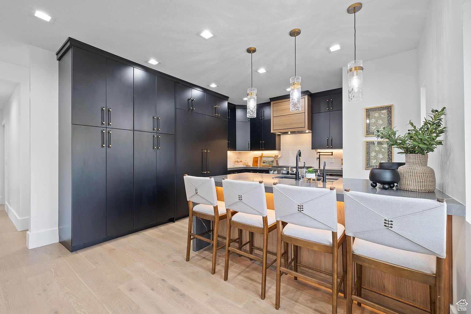 This is a modern kitchen featuring sleek, dark cabinetry with gold hardware, complemented by light wood flooring. A central island with bar seating is illuminated by pendant lights, and decorative items add a touch of elegance. The kitchen presents a clean and sophisticated aesthetic.