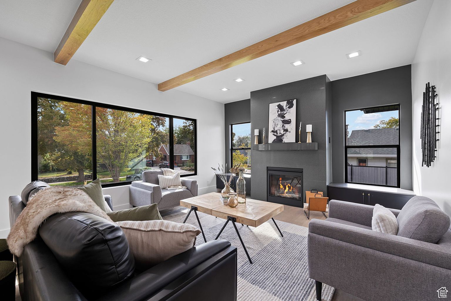 This is a modern living room featuring a gray color scheme with wooden beam accents on the ceiling. The room has a fireplace with a dark gray tile surround and a contemporary art piece above it. Large windows provide natural light and views of the outside, while comfortable seating arrangements invite relaxation.