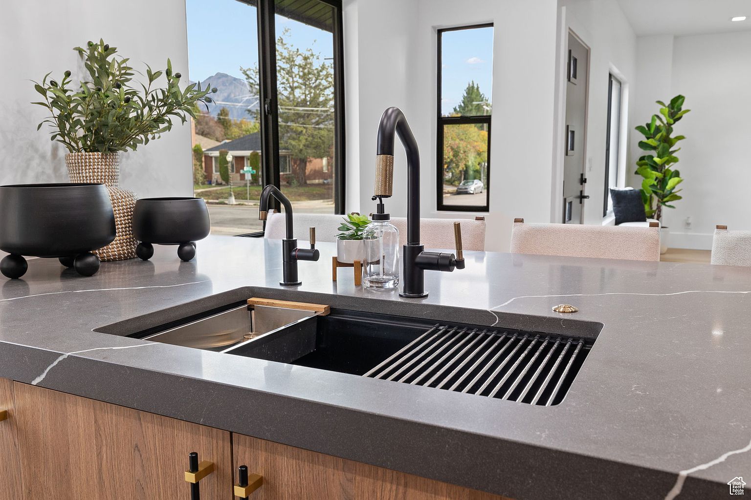 This image showcases a modern kitchen island with a dark gray countertop and a unique sink design featuring a built-in grate. The island is adorned with sleek black fixtures, including a faucet and soap dispenser, complemented by decorative plants and stylish black bowls. The kitchen's design is contemporary, with a focus on clean lines and sophisticated details.