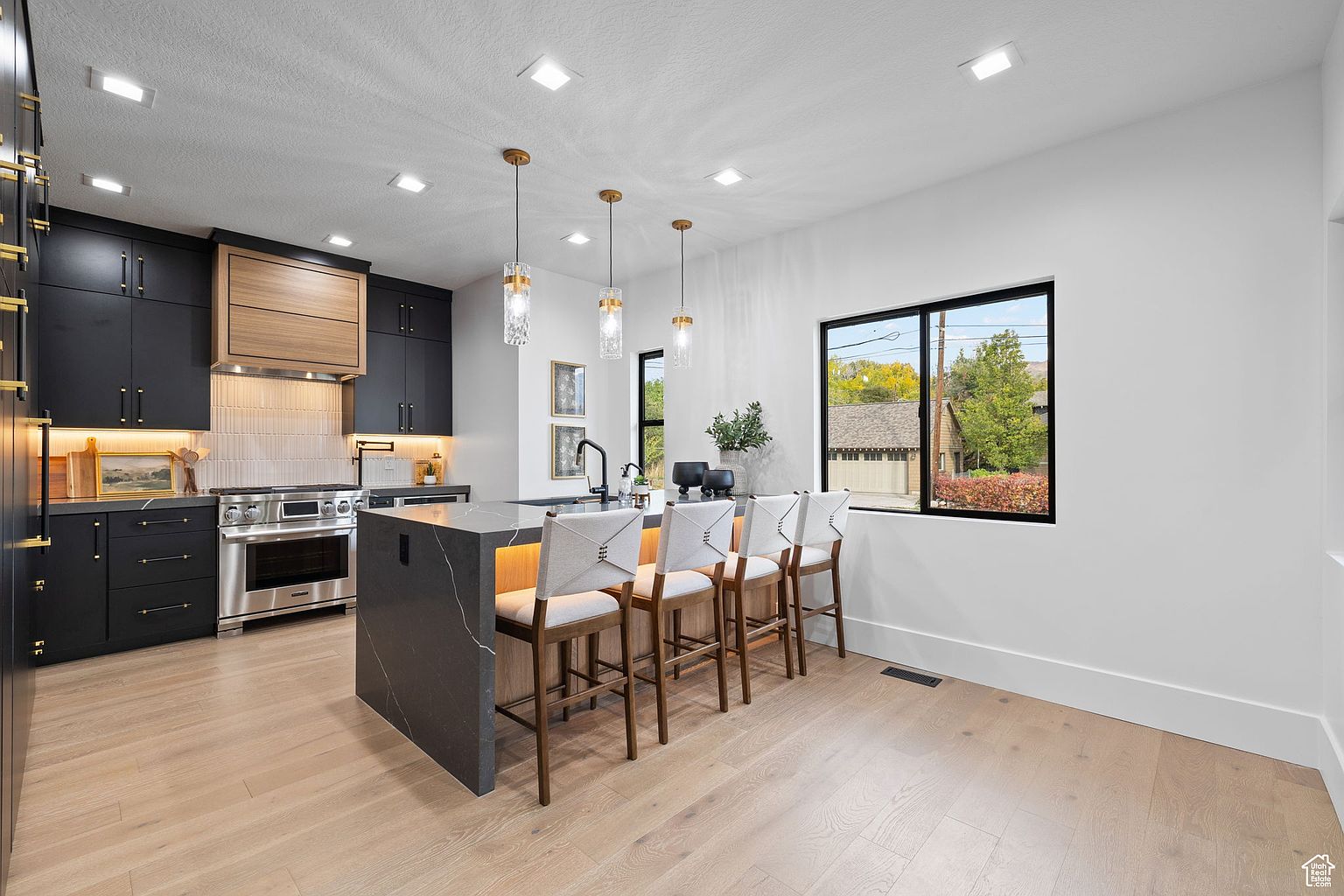 This is a modern kitchen with dark cabinetry accented by gold hardware and under-cabinet lighting. A large island with a waterfall countertop provides seating for four, illuminated by pendant lights. The kitchen features stainless steel appliances and a window offering a view of the outdoors, creating a bright and inviting space.