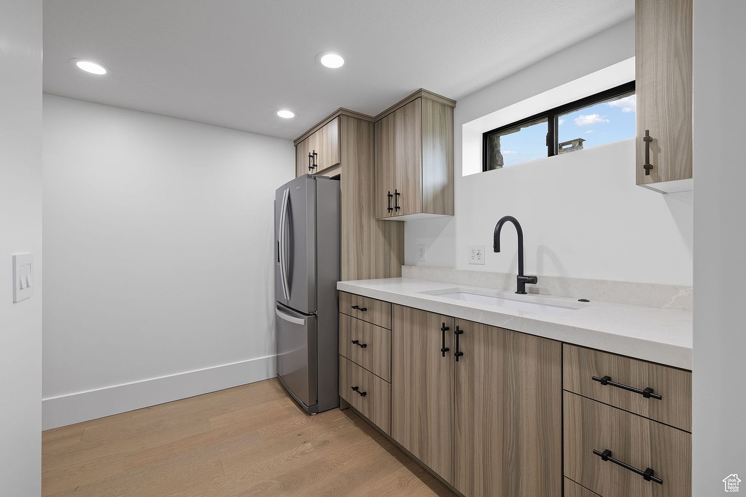 This is a well-lit kitchen featuring light wood-grain cabinets with black hardware and a white countertop. A stainless steel refrigerator stands to the left, and a black faucet is installed in the sink. The flooring is light wood, and the walls are painted white, creating a clean and modern aesthetic.