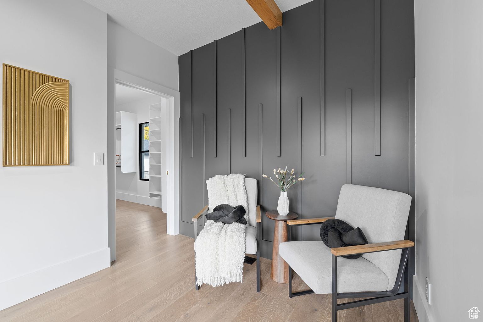 This interior shot showcases a stylish hallway or landing area with modern design elements. Two chairs, one draped with a cozy throw, flank a small side table with a vase of flowers, set against a dark gray paneled wall. A gold artwork hangs on the adjacent white wall, and a glimpse into a bright, organized closet is visible through an open doorway, creating an inviting and sophisticated atmosphere.