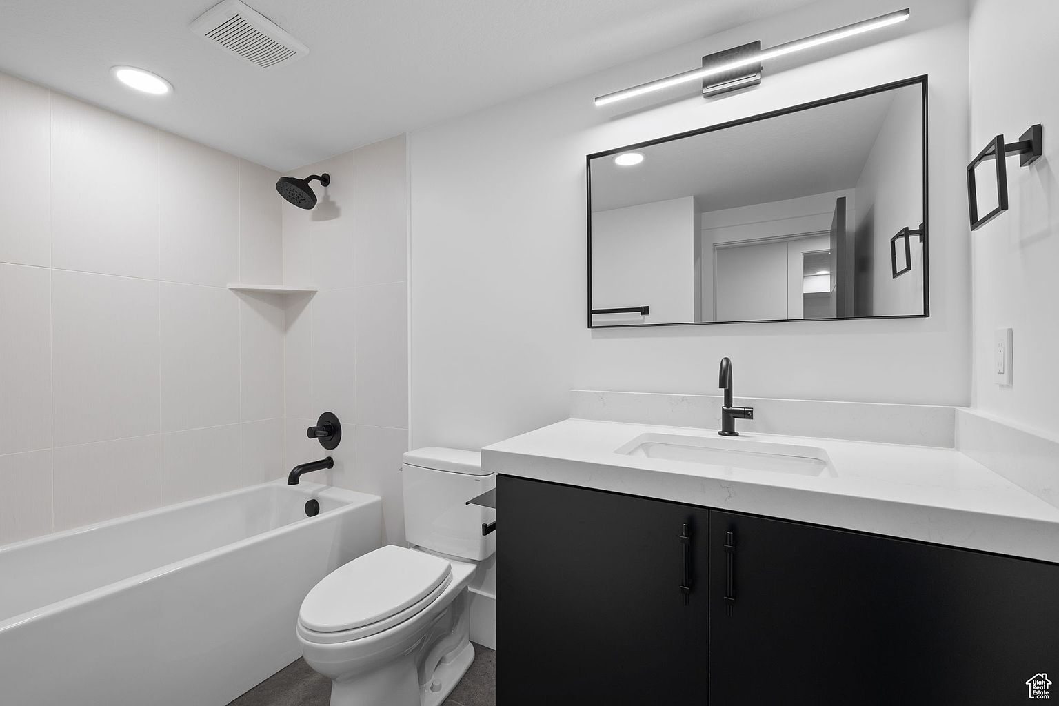 This is a well-lit bathroom featuring a modern design with a black and white color scheme. The bathroom includes a bathtub with a shower, a toilet, and a vanity with a white countertop and black cabinets. A large mirror hangs above the vanity, reflecting the clean and minimalist aesthetic of the space.