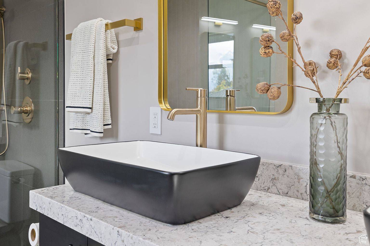 This is a close-up shot of a modern bathroom vanity. The vanity features a black rectangular vessel sink with a white interior, a gold-toned faucet, and a light-colored countertop with subtle veining. A gold-framed mirror hangs above the sink, and a decorative vase with dried floral elements sits to the side, adding a touch of elegance.