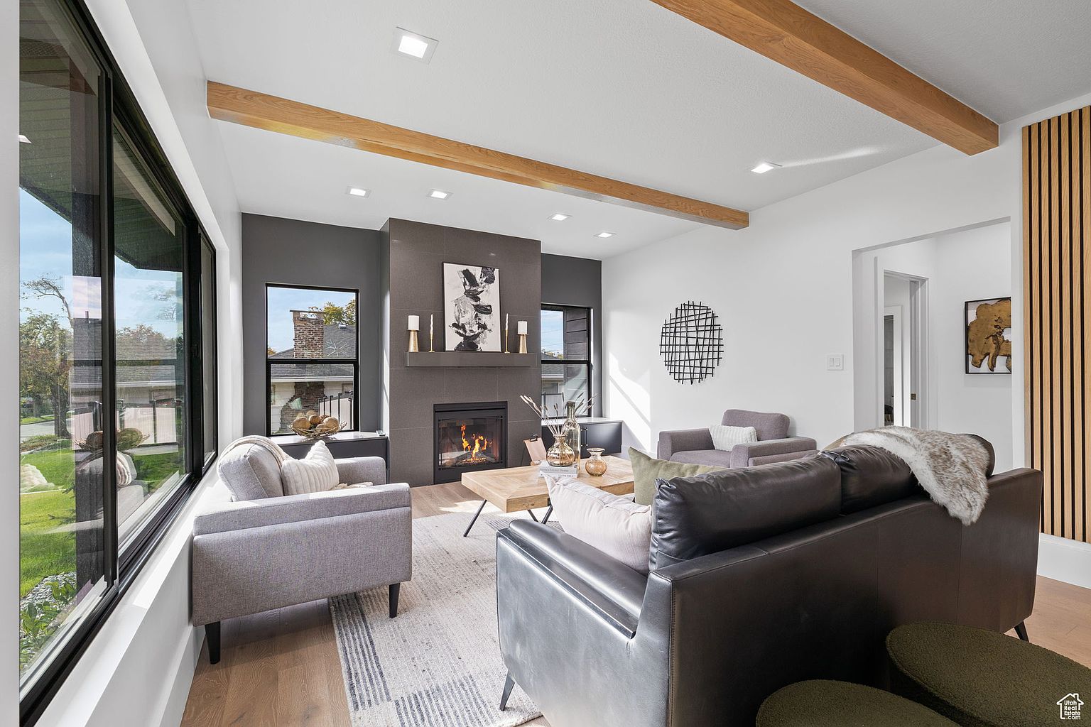 This is an interior shot of a modern living room. The room features a fireplace with a dark gray surround, wooden ceiling beams, and large windows that provide ample natural light. The furniture includes a gray sofa, a leather sofa, and a light-colored area rug, creating a comfortable and stylish living space.