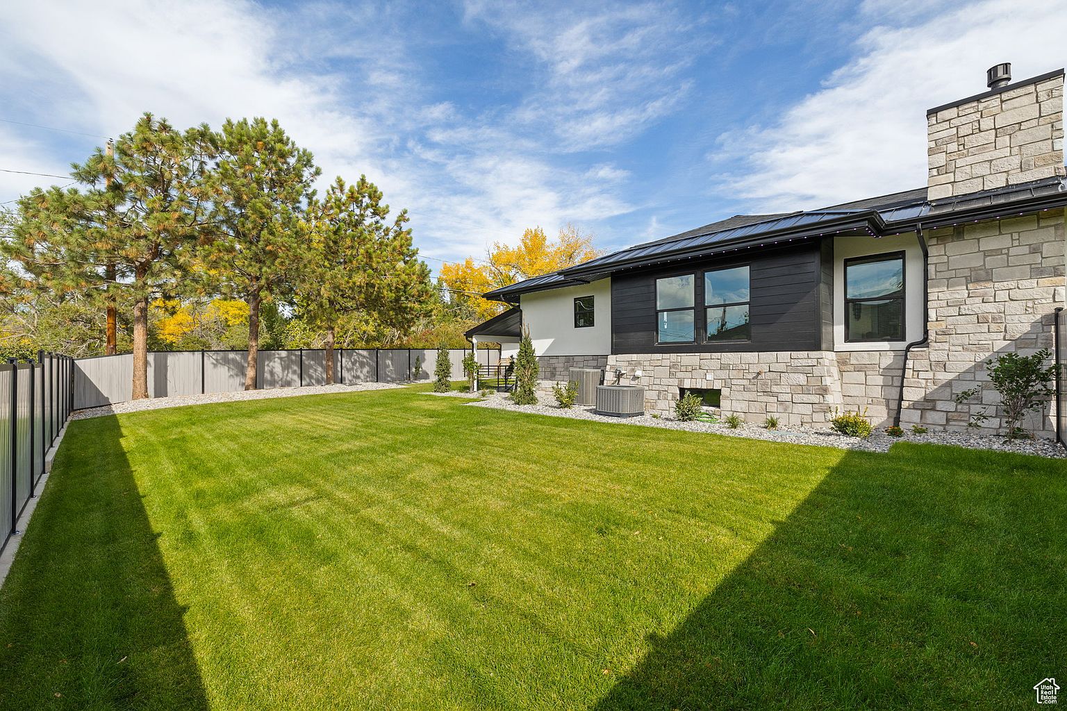 This image showcases a well-maintained backyard featuring a lush green lawn, mature trees, and a modern home exterior. The house has a combination of stone and dark siding, with large windows providing ample natural light. A fence encloses the yard, offering privacy and security, while the overall impression is one of tranquility and upscale living.