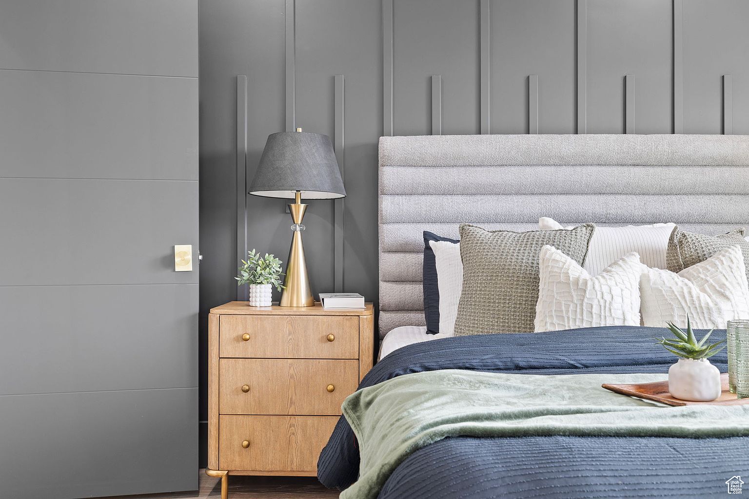 This is a well-staged primary bedroom featuring a gray upholstered headboard, a nightstand with a lamp and decorative plant, and a neatly made bed with multiple pillows and a throw blanket. The color palette is soothing, with shades of gray, blue, and green, creating a relaxing and inviting atmosphere. The room showcases modern design elements and attention to detail, making it an appealing space for potential buyers.