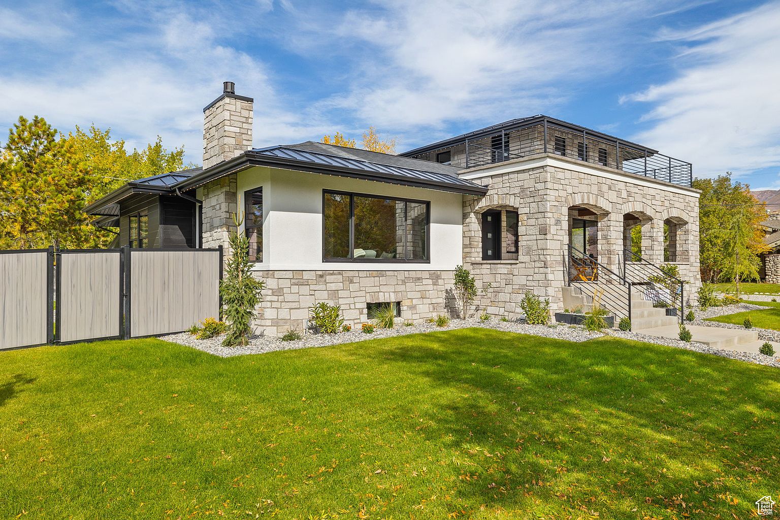 This is a front exterior view of a modern stone house with a well-manicured lawn. The house features a combination of stone and stucco, with a black metal roof and window frames. A stone chimney adds character, and a fenced yard provides privacy. The overall impression is one of luxury and curb appeal.