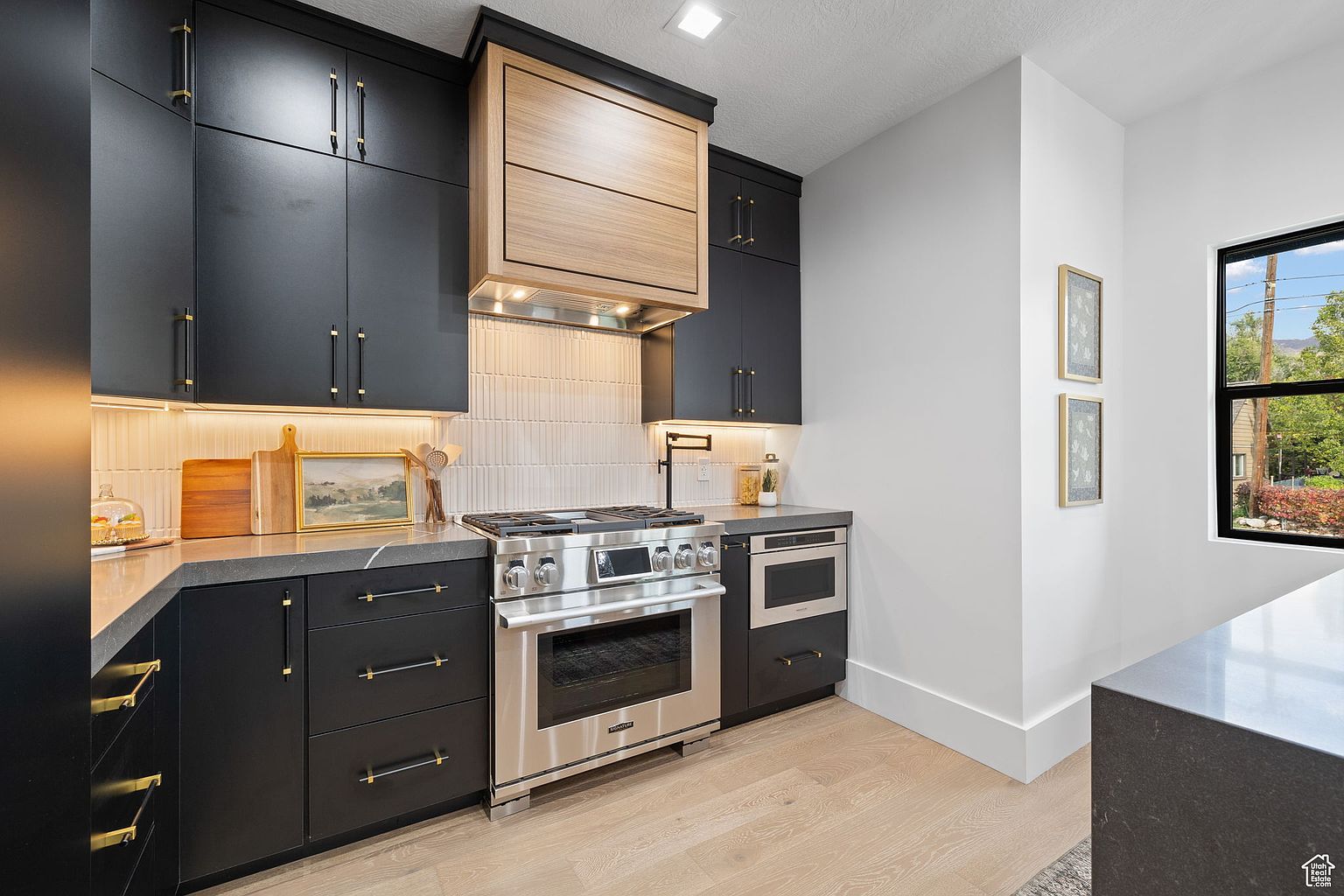 This is a modern kitchen featuring sleek, black cabinetry with gold hardware, complemented by a stainless steel range and oven. A light wood accent above the range adds warmth, while the white backsplash and walls create a bright contrast. The hardwood flooring and natural light from the window enhance the contemporary design.