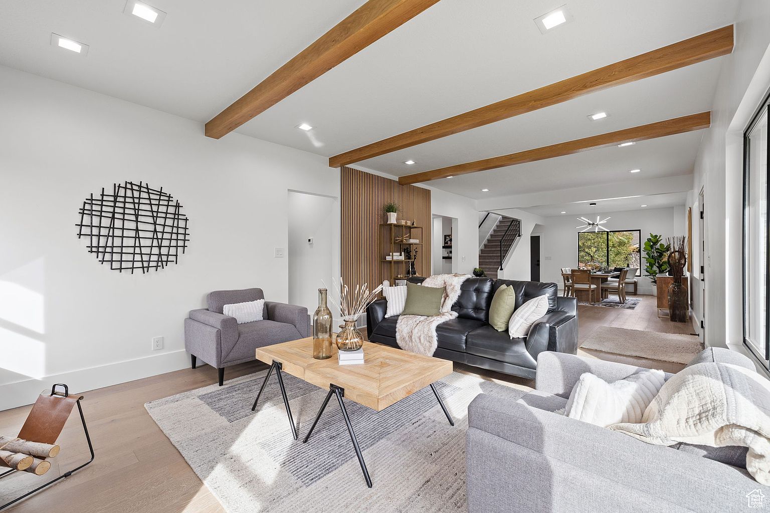 This is a bright and airy living room featuring hardwood floors, white walls, and exposed wooden beams on the ceiling. The room is furnished with a black leather sofa, two gray armchairs, and a square wooden coffee table. A modern, circular wall art piece adds a touch of sophistication, while the open floor plan leads to a dining area, creating a seamless flow.
