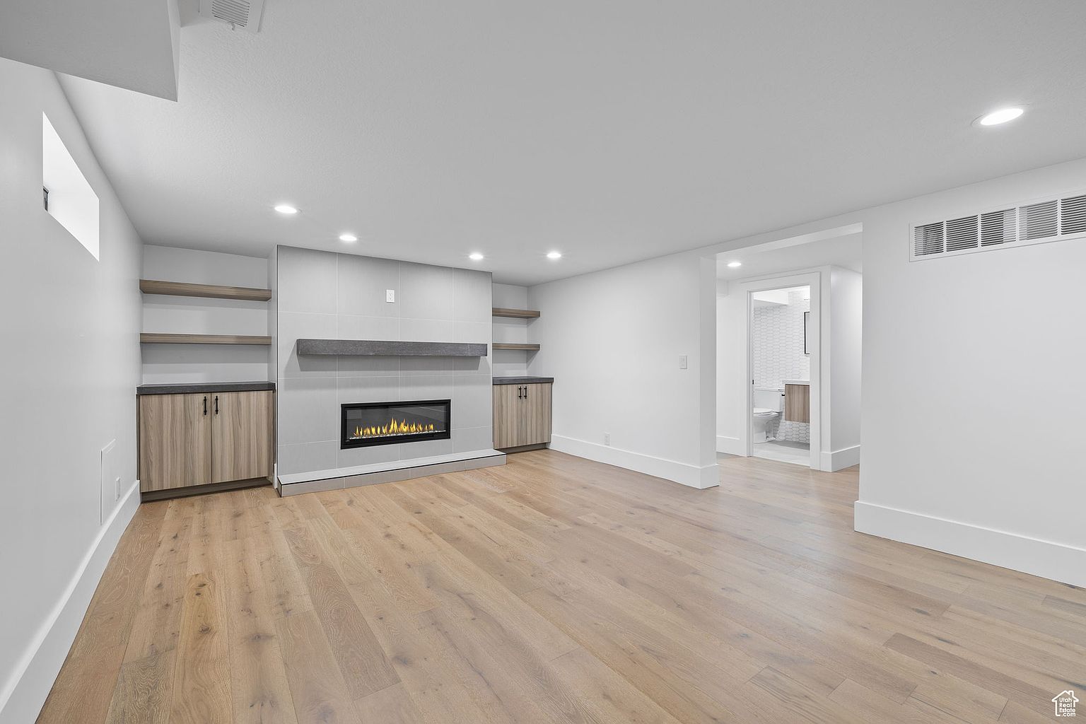 This is a bright and modern living room featuring light wood flooring and white walls. A fireplace with a gray mantle is the focal point, flanked by built-in cabinets and shelves. A doorway leads to a bathroom, and the overall impression is clean and spacious.