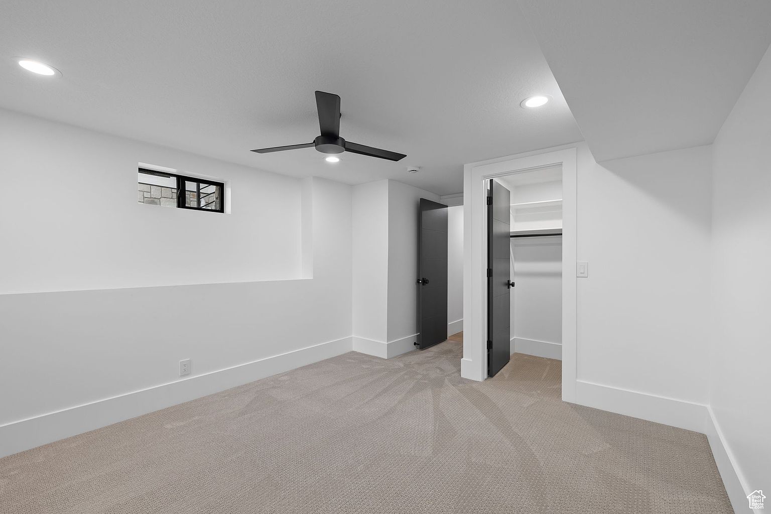 This is a clean and modern bedroom with white walls, light gray carpet, and a black ceiling fan. The room features a small window, a closet, and a doorway leading to another room. The overall impression is minimalist and bright, suitable for a guest bedroom or a secondary bedroom in a home.