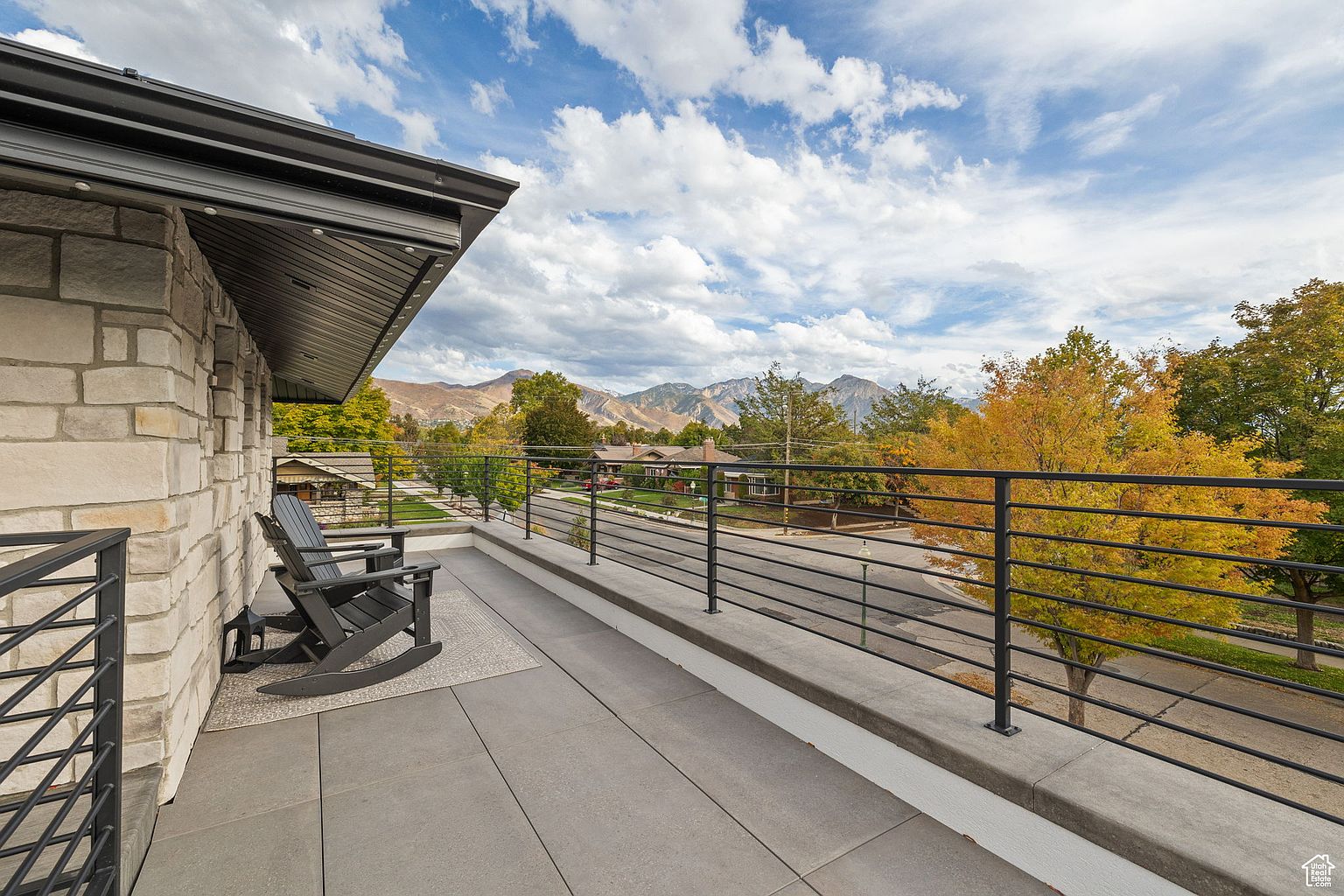 This image showcases a spacious balcony with a modern design, featuring gray tiled flooring and a sleek black railing. Two dark-colored rocking chairs are positioned on a patterned rug, inviting relaxation. The balcony offers a scenic view of the neighborhood, mountains, and a partly cloudy sky, enhancing the property's appeal.