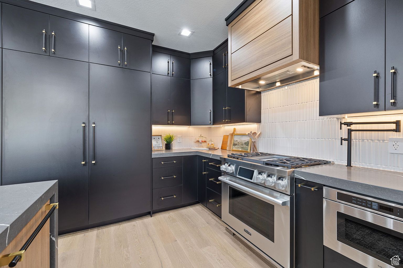 This is a modern kitchen with dark cabinetry and gold hardware. Stainless steel appliances are present, including a range and oven. The countertops are a light gray, and the backsplash is a textured white tile. The flooring is light wood, and the overall style is sleek and contemporary.