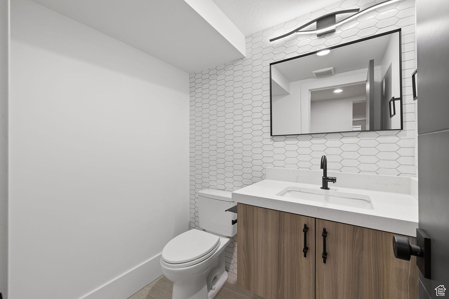 This is a well-lit guest bathroom featuring a modern design. The space includes a toilet, a vanity with a white countertop and wood-look cabinetry, a black faucet, and a rectangular mirror with a modern light fixture above. The wall behind the vanity is adorned with a geometric tile pattern, while the other walls are painted white, creating a clean and contemporary aesthetic.