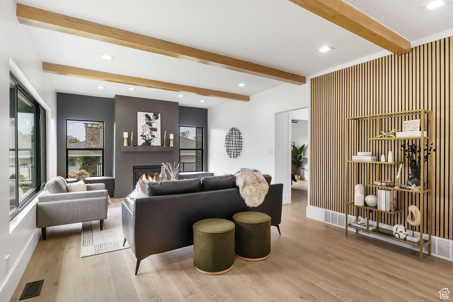 This is a stylish living room featuring a modern fireplace with a gray surround and a contemporary art piece above. The room is decorated with wooden ceiling beams, a dark leather sofa, and olive green ottomans. A decorative wooden slat wall with a gold shelving unit adds visual interest, and the hardwood floors contribute to the room's warm and inviting atmosphere.