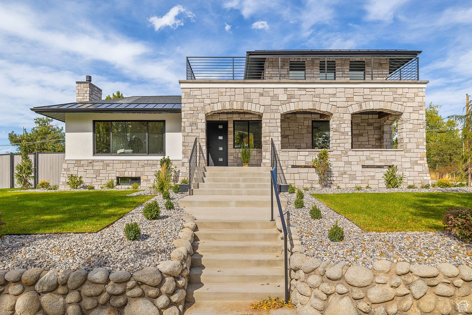 This is a front view of a modern two-story house with a stone facade. The house features a flat roof with a balcony, arched entryways, and a well-manicured lawn. A stone retaining wall and concrete steps lead to the front door, creating an inviting entrance.