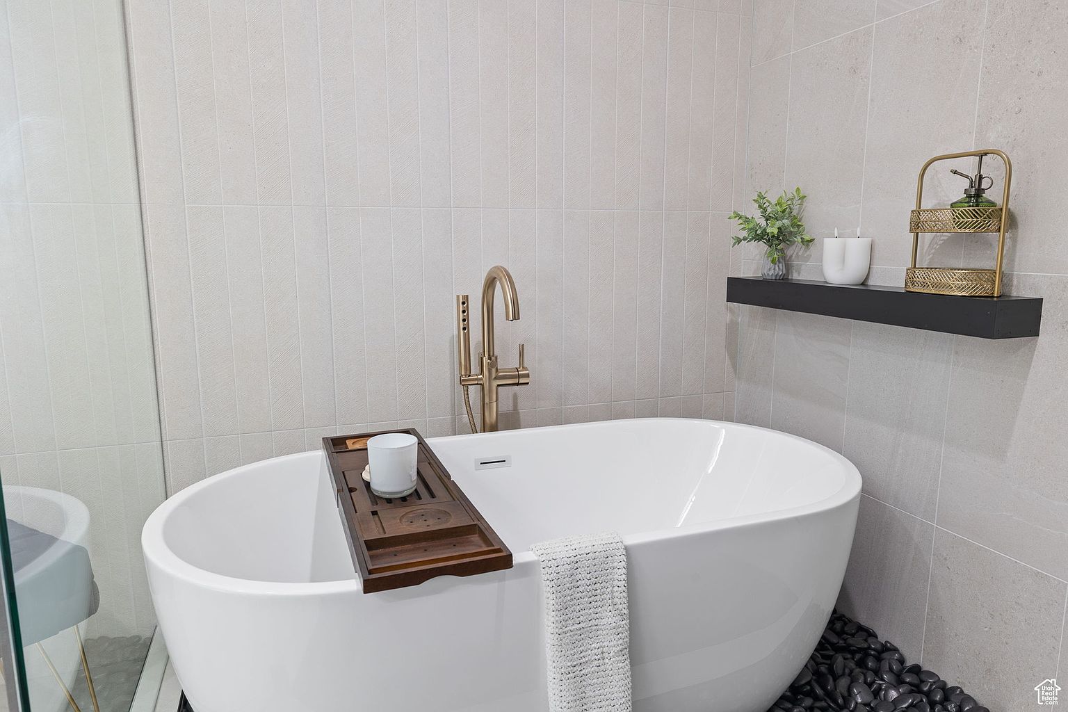 This is a well-appointed primary bathroom featuring a freestanding white bathtub with a gold faucet and a wooden bath tray holding a candle. A black shelf displays decorative items, and the walls are tiled in a neutral tone. The overall impression is one of luxury and relaxation.