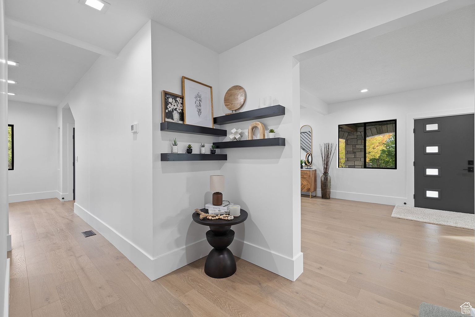 This interior shot showcases a modern hallway with light wood flooring and white walls. The hallway features decorative floating shelves with artwork and plants, adding a touch of elegance. A dark wood side table with a lamp and decorative items sits in the corner, enhancing the stylish and welcoming atmosphere.