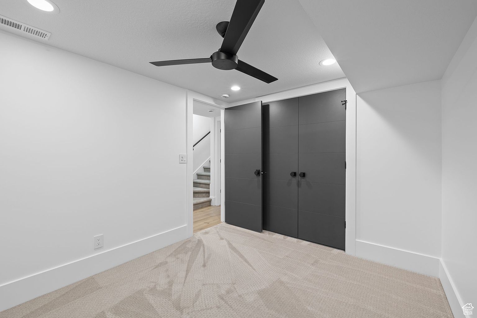 This is a well-lit bedroom featuring neutral-toned carpet and white walls, creating a bright and airy atmosphere. A modern black ceiling fan adds a stylish touch, while dark gray closet doors provide ample storage. An open doorway reveals a staircase, suggesting access to another level of the home.