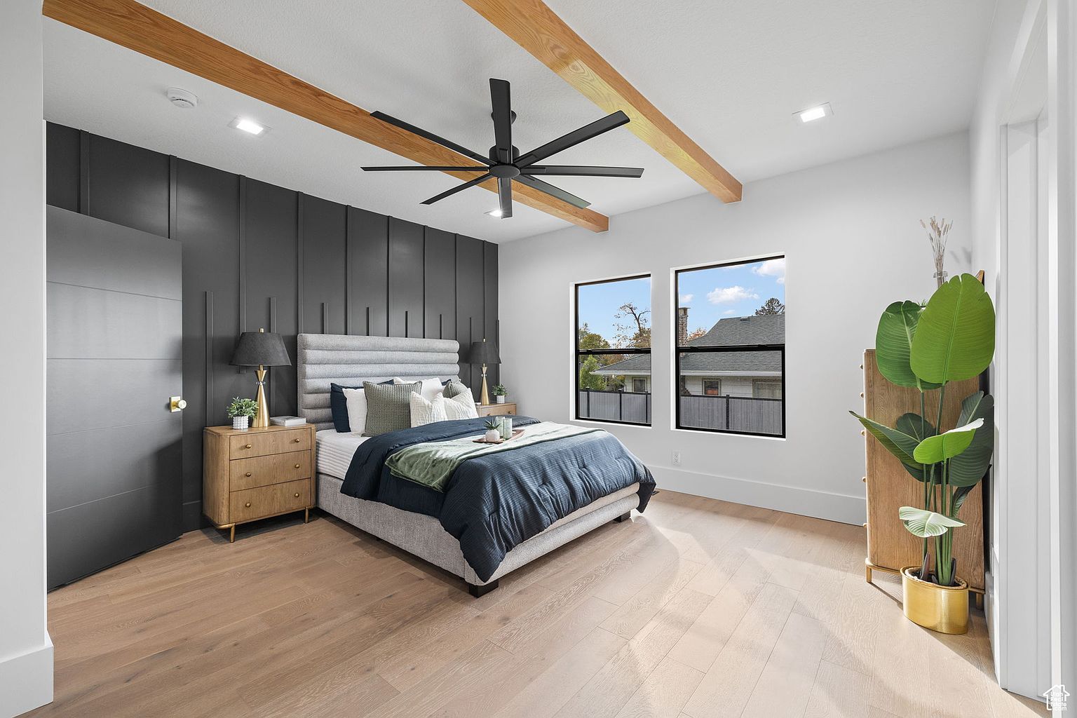 This is a beautifully staged primary bedroom featuring a modern design with a dark accent wall and light wood flooring. The room includes a large bed with a padded headboard, two windows providing natural light, and wooden ceiling beams adding a touch of rustic charm. A large houseplant adds a touch of nature to the space.