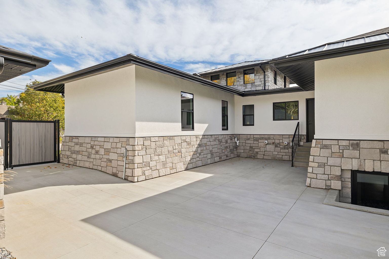 This is a side view of a modern home featuring a combination of white stucco and stone veneer on the exterior. The design incorporates clean lines, dark-framed windows, and a dark roof, creating a contemporary aesthetic. A concrete driveway leads to a gated area, enhancing the property's curb appeal.