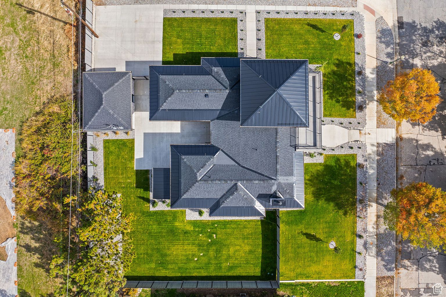 This aerial shot showcases a modern home with a dark gray metal roof, meticulously landscaped grounds, and a concrete driveway. The property features well-manicured lawns, decorative gravel borders, and mature trees, creating a visually appealing and upscale impression. The perspective highlights the architectural design and the overall layout of the property within its surroundings.