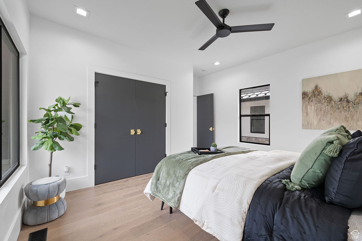 This is a well-lit primary bedroom featuring a modern aesthetic. The room includes a bed with a black, white, and green color scheme, complemented by a black ceiling fan and gray closet doors with gold hardware. A potted plant sits near the window, adding a touch of nature to the space.