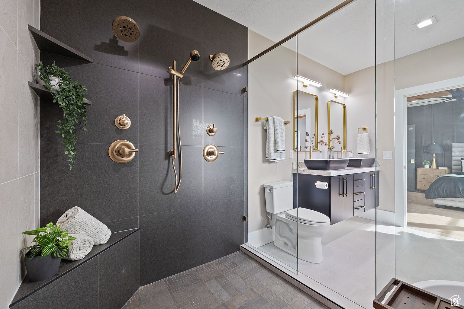 This is a modern primary bathroom featuring a walk-in shower with dual shower heads and bronze fixtures, a toilet, and a double vanity with black vessel sinks. The shower has dark gray tiling, while the rest of the bathroom has light gray tiling. The vanity is dark blue with bronze hardware, and the mirrors are framed in bronze. The overall impression is luxurious and contemporary.