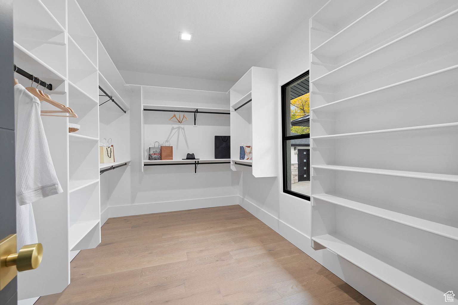 This is a bright and spacious walk-in closet featuring custom white shelving and black hanging rods. The closet has a light wood floor and a window providing natural light. The overall impression is clean, organized, and luxurious, offering ample storage space.