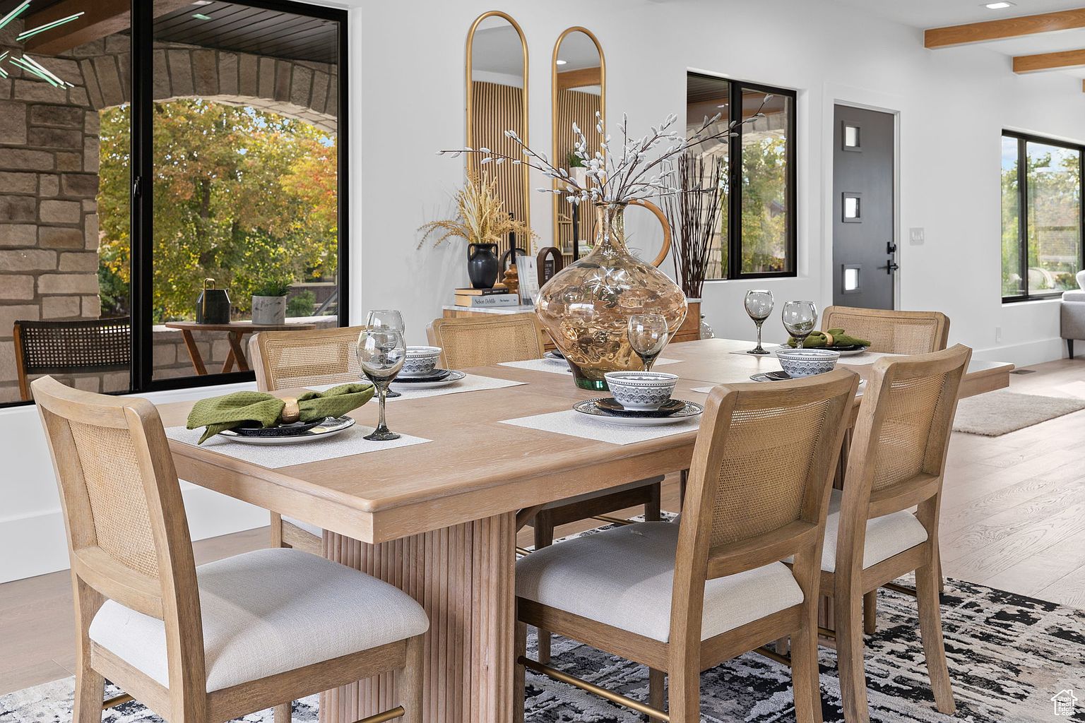 This is an interior shot of a dining room featuring a large wooden table set for a meal. The table is surrounded by six chairs with light-colored cushions and woven backs. A large decorative vase sits in the center of the table, and natural light streams in through the windows, creating a warm and inviting atmosphere.