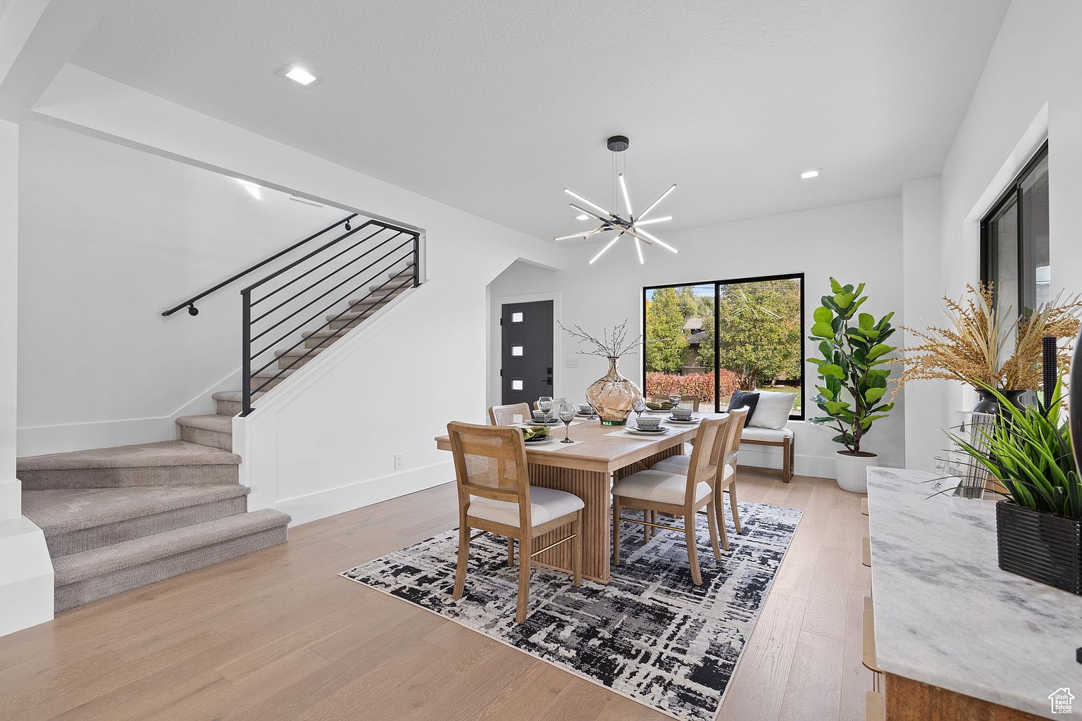 This is an interior shot of a dining room featuring a wooden dining table with four chairs, set on a black and white patterned rug. A modern chandelier hangs above the table, and a large window provides natural light. The room also includes a staircase with black railings and a decorative plant, creating a bright and inviting space.