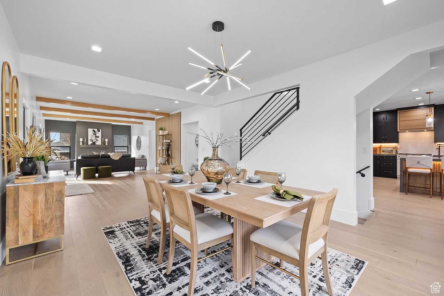 This is an interior shot of a dining room featuring a light wood dining table set for six, with modern chairs and a patterned rug underneath. A contemporary chandelier hangs above the table, and the room opens to a living area and a staircase, creating an open and airy feel. The perspective is from a wide angle, showcasing the dining area's connection to the rest of the home.