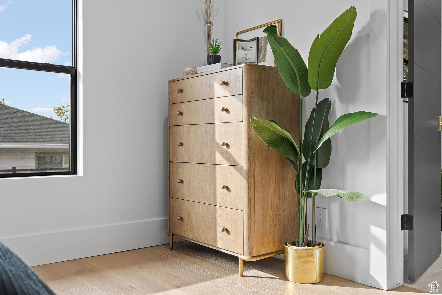 The image showcases a well-lit bedroom interior featuring a light wood dresser with gold hardware, complemented by a tall potted plant in a gold pot. Natural light streams in from a nearby window, highlighting the hardwood flooring and creating a warm, inviting atmosphere. The room's design is simple yet elegant, emphasizing natural materials and a clean aesthetic.