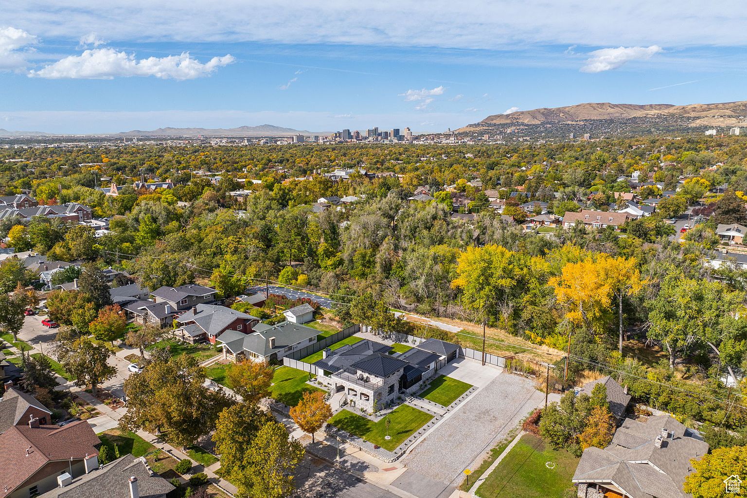 This aerial shot showcases a modern home with a dark roof, light stone facade, and well-manicured lawn. The property features a large paved driveway and is surrounded by mature trees, offering privacy. In the background, a cityscape and mountains add to the scenic view, suggesting a desirable location.