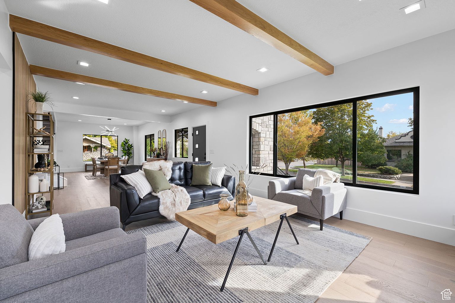 This is a bright and airy living room featuring a black leather sofa, gray armchairs, and a light wood coffee table on a striped rug. The room has wooden ceiling beams and a large window with a view of the outdoors, creating a modern and inviting space. A decorative shelving unit is visible in the background, adding to the room's character.